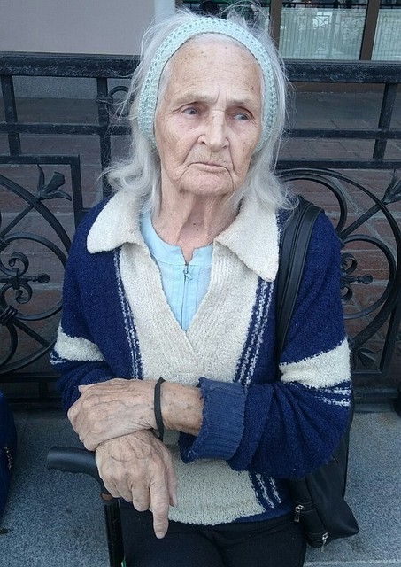 Бабушка одета в черные брюки, синюю кофту, с повязкой на седых волосах и крюкой